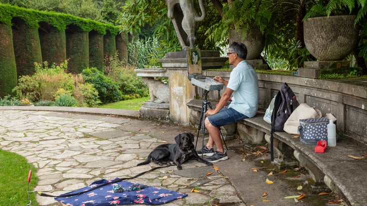 A man sitting on a bench painting in the garden at Mount Stewart, while his dog lies on the ground beside him